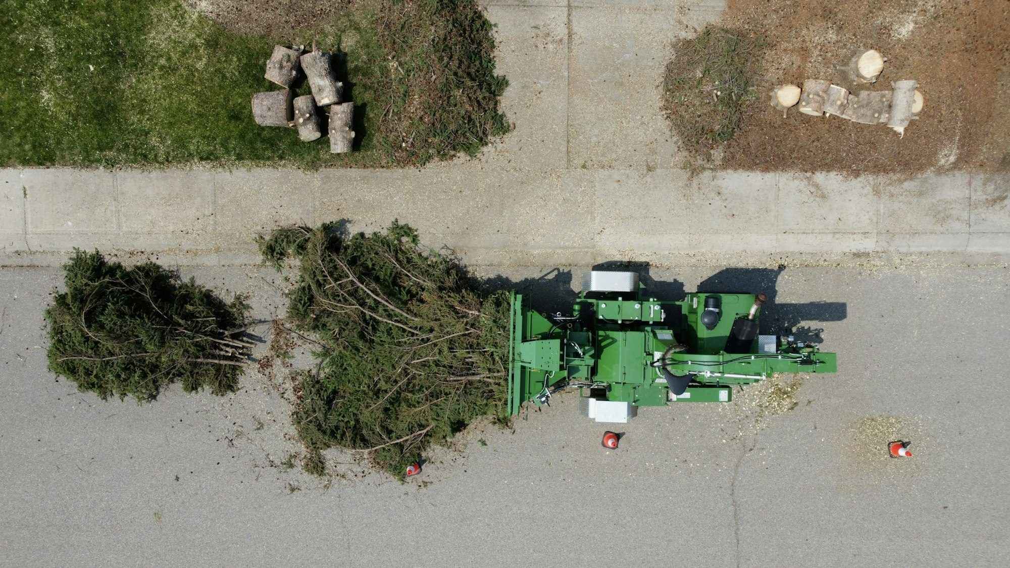 Overhead view of a green wood chipper on a suburban street beside piles of cut logs and evergreen branches, with orange traffic cones