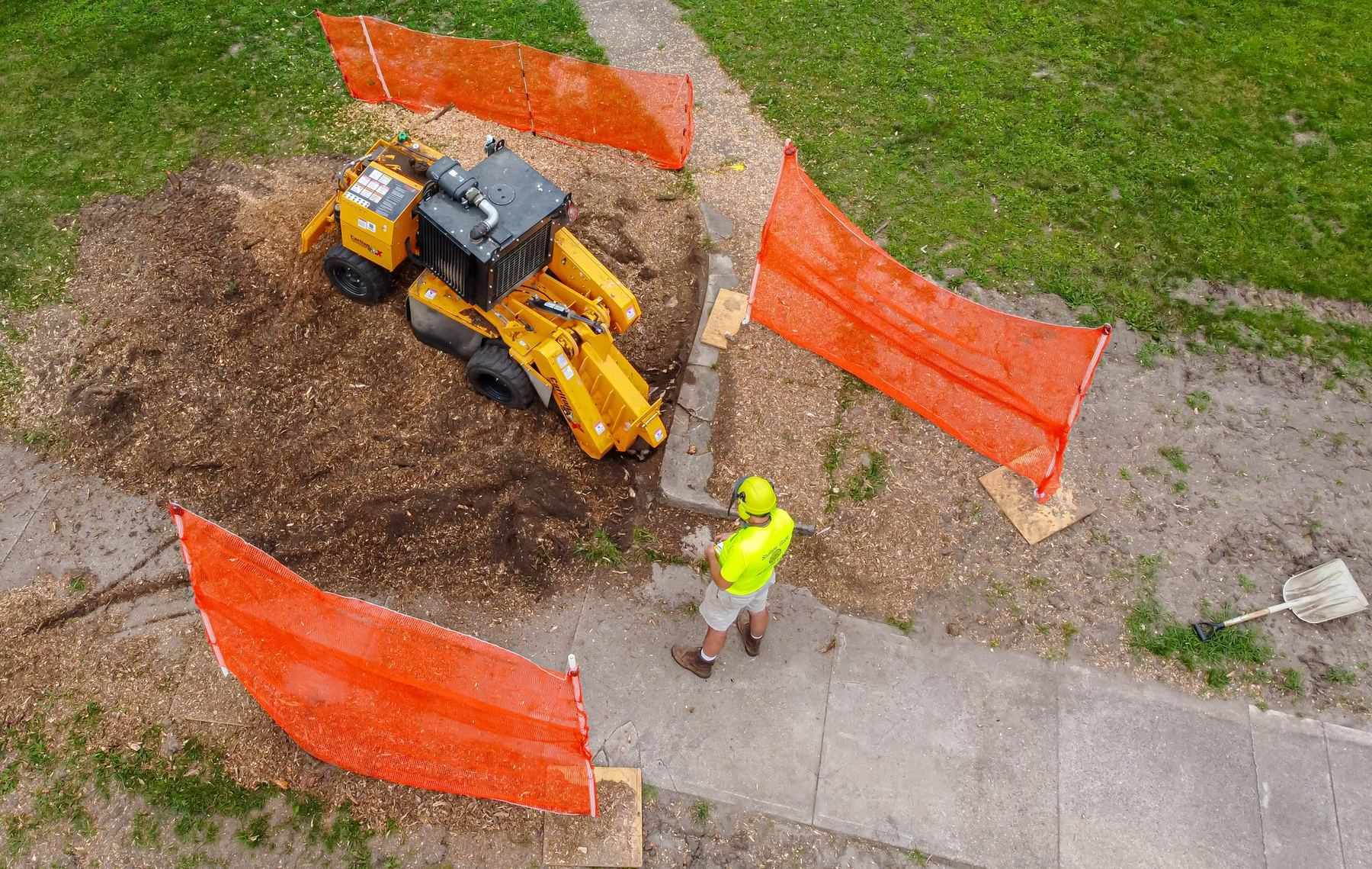 Yellow tracked stump grinder working on a suburban lawn beside orange mesh safety screens, with a worker in a hi-vis shirt and hard hat on the path
